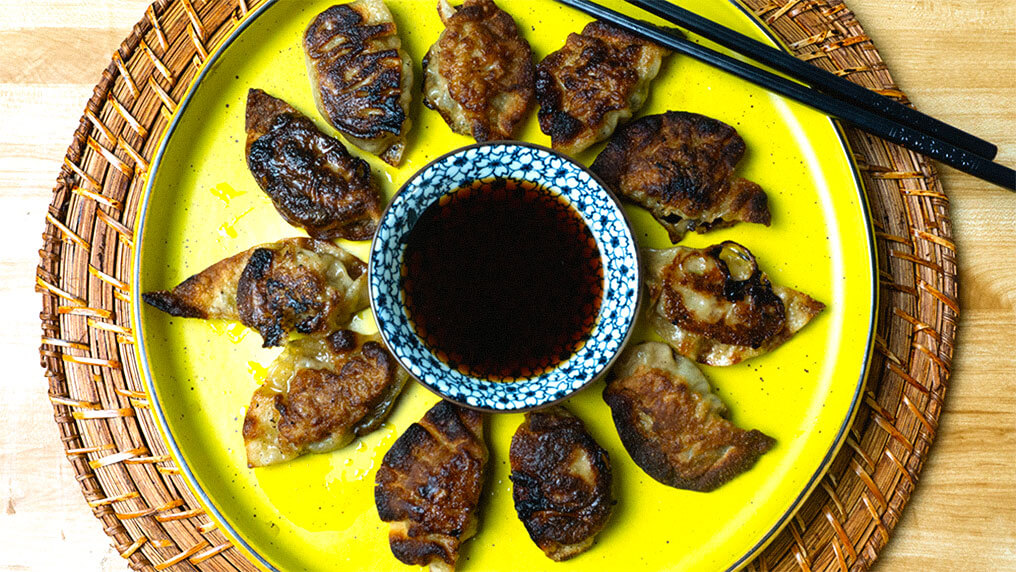 gyoza on yellow plate, with chocksticks on the right side with a soysauce asian blue bowl in the center