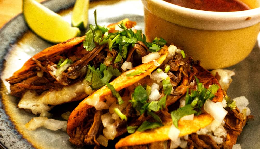 Birria tacos topped with cillantro and onions with lime wedges and consume.